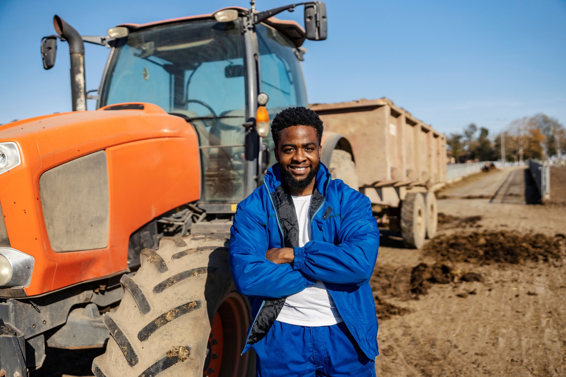 Portrait of black farmer standing next to tractor at farmyard and smiling at camera.